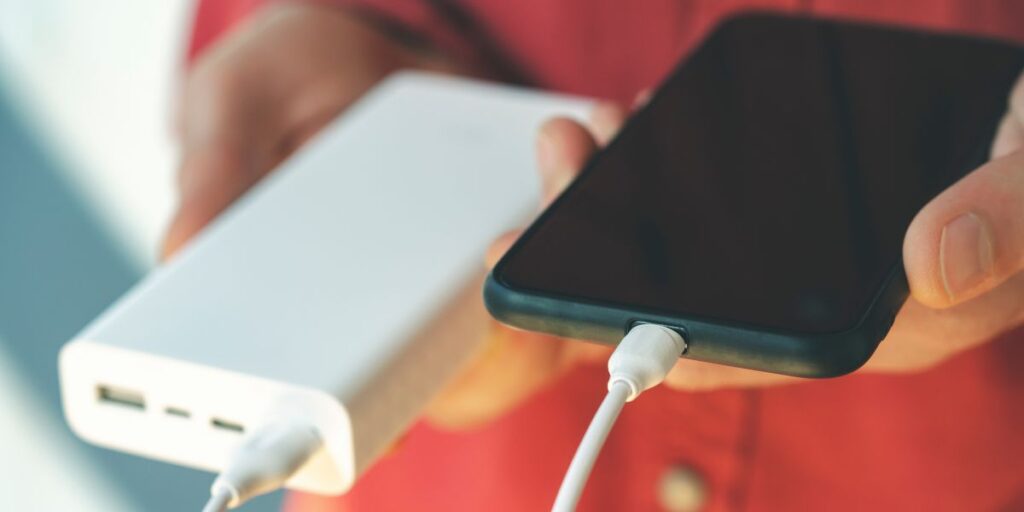 A close-up of hands connecting a white power bank to a smartphone with a charging cable, indicating portable charging on the go.