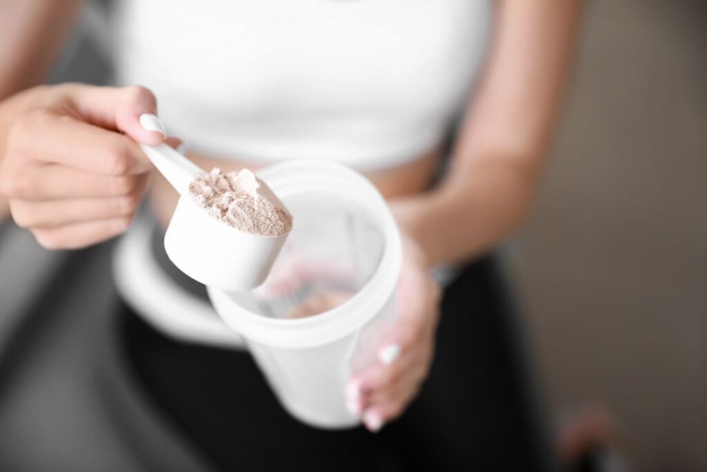 The image shows a person scooping protein powder from a container with a white measuring scoop. The background features a blurred figure in a white tank top, suggesting a fitness or health-related setting.