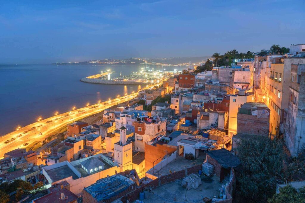 Tangier, Morocco, a coastal city at dusk with tightly packed hillside buildings and a brightly lit seaside road curving along the water toward a glowing harbor.