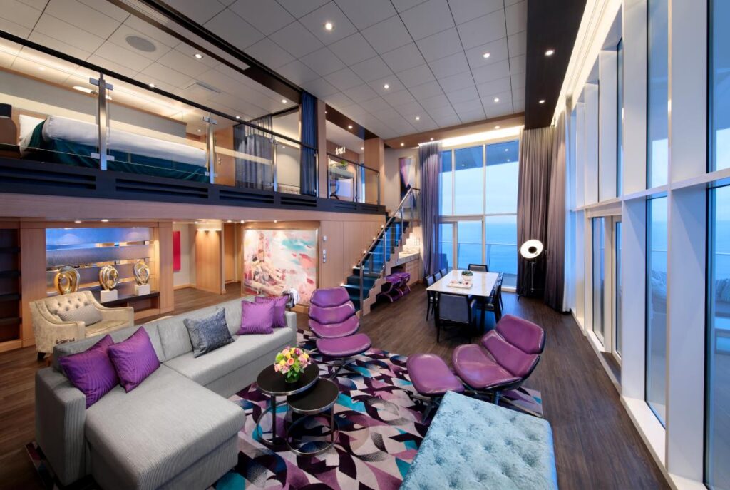 Luxurious two-level Royal Loft Suite on Harmony of the Seas featuring a spacious living area with modern furnishings, vibrant purple accents, and floor-to-ceiling windows offering an expansive ocean view.