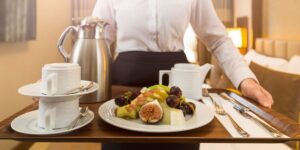 A server in a crisp white shirt presents a hotel room service tray with a variety of fresh fruits on a plate, including slices of fig, apple, and grapes, accompanied by stacked white coffee cups and a silver coffee pot, suggesting a luxurious and convenient dining experience.