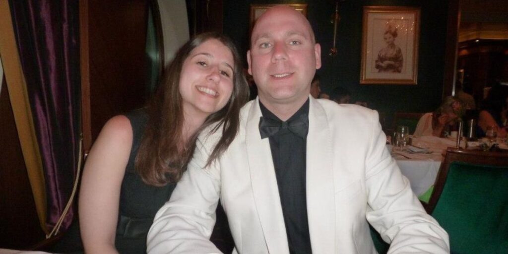 A smiling couple dressed in evening attire seated at a table in a sophisticated dining room, with a classic portrait and other diners in the background, capturing a moment of a Royal Caribbean dining experience.