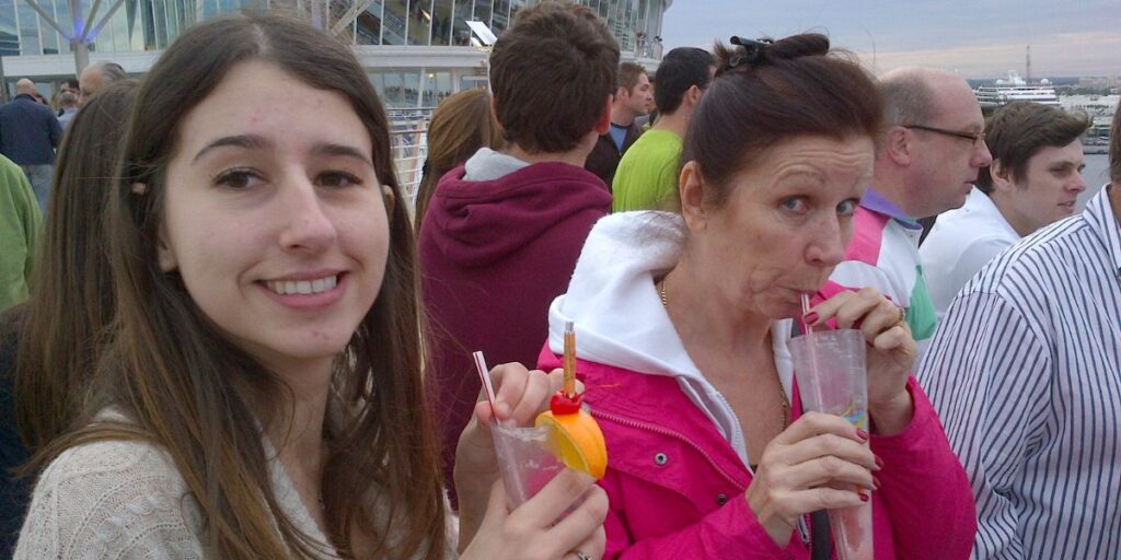 Two women enjoying tropical drinks on the deck of a Royal Caribbean cruise ship, with a bustling crowd and the ship's structure in the background. The younger woman smiles at the camera while the other sips her drink with a playful expression.