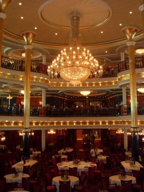 The main dining room on a Royal Caribbean cruise ship, featuring a grand chandelier as the centerpiece, with multiple levels of balconied seating. The elegantly set tables and warm lighting create a luxurious dining experience.