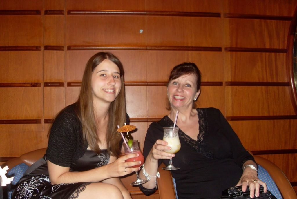 Two women enjoying cocktails on Royal Caribbean cruise, the younger with a strawberry daiquiri and the elder with a pina colada, seated in a lounge with wooden panels, radiating warmth and leisure.