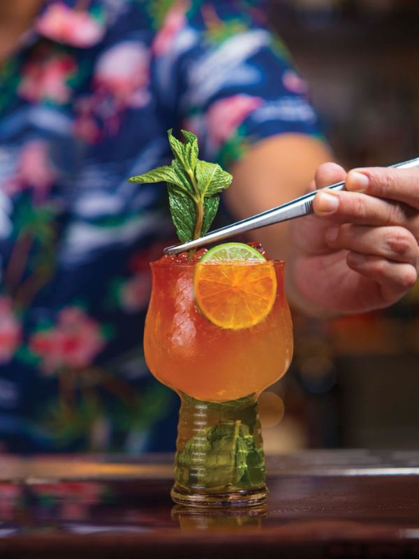 A tropical cocktail being served on a Royal Caribbean cruise, garnished with a fresh sprig of mint and a slice of orange, presented in a uniquely textured tiki glass, suggesting an inviting and refreshing beverage experience.