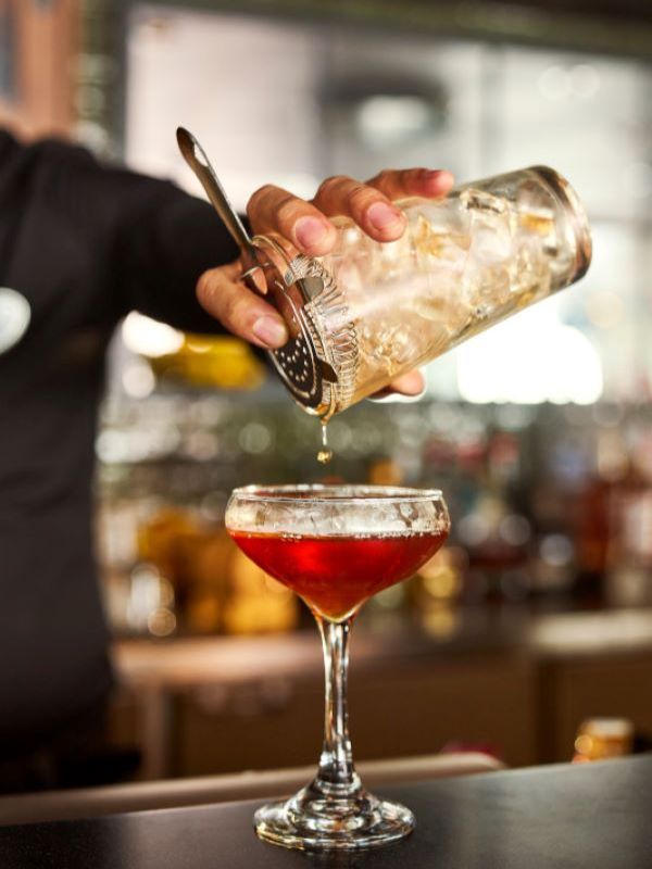 Bartender straining a red cocktail into a coupe glass at a bar, with the background softly blurred.
