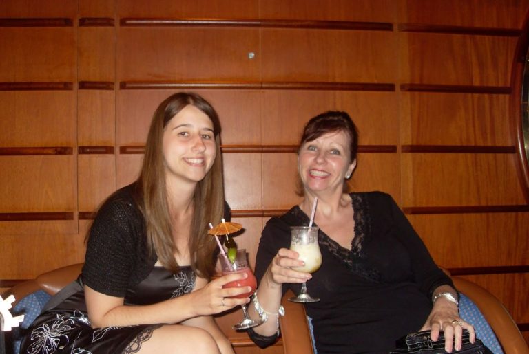 Two women enjoying cocktails on Royal Caribbean cruise, the younger with a strawberry daiquiri and the elder with a pina colada, seated in a lounge with wooden panels, radiating warmth and leisure.