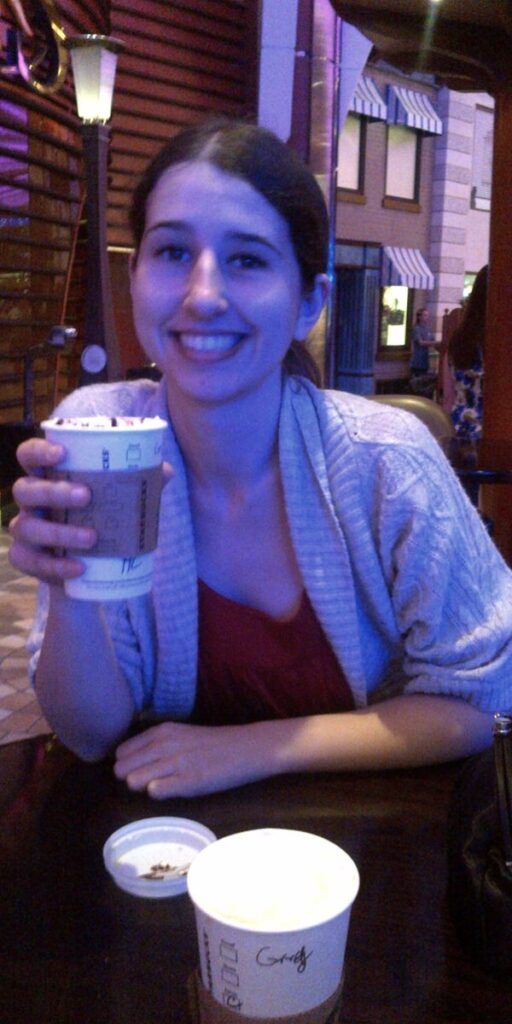 A smiling woman holding a coffee cup sits in a cozy, dimly lit interior of a Royal Caribbean cruise ship café, with ambient lighting and part of the ship's promenade visible in the background.
