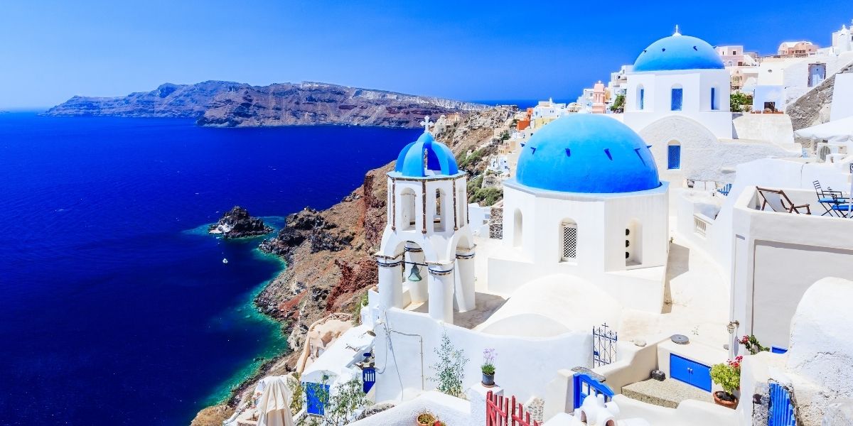 A picturesque view of Santorini, Greece, featuring iconic whitewashed buildings with vibrant blue domes overlooking the deep blue Aegean Sea. The cliffsides are dotted with pastel-colored houses, and the rugged coastline stretches into the distance under a clear, sunny sky.