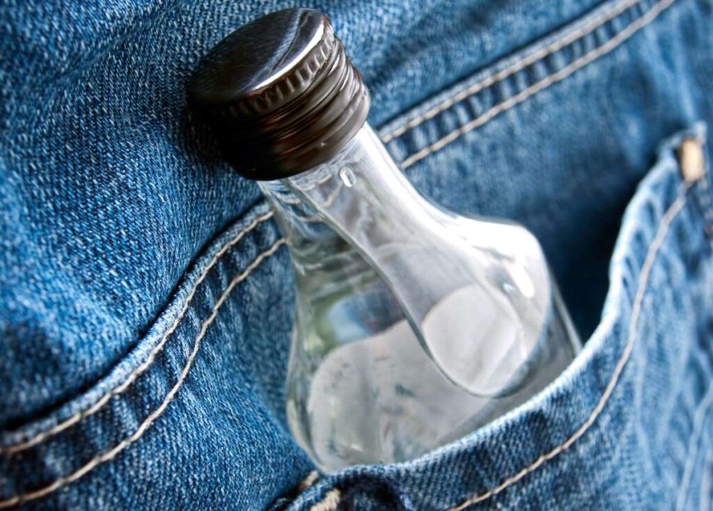 Close-up of a clear glass flask with a black cap tucked into the pocket of blue denim jeans, conceptually highlighting discreet transportation of alcohol.