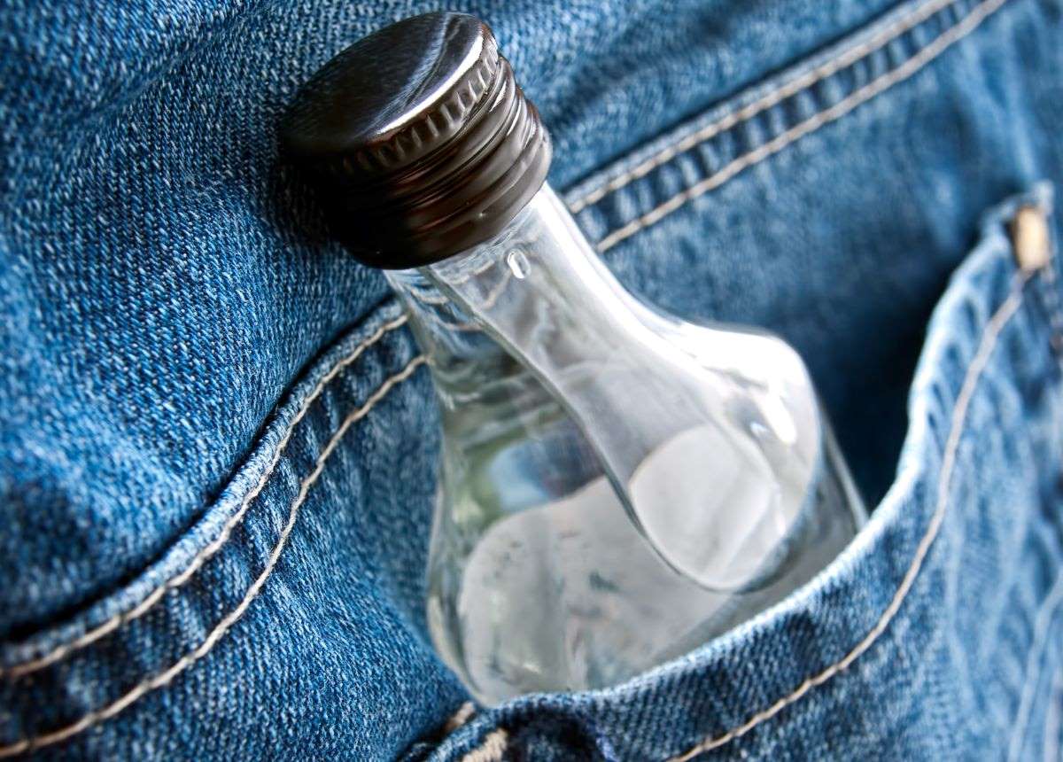 Close-up of a clear glass flask with a black cap tucked into the pocket of blue denim jeans, conceptually highlighting discreet transportation of alcohol.