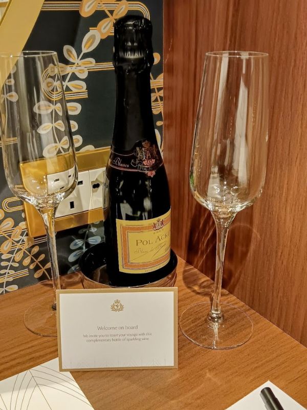 The image shows a welcoming setup inside a cabin on Cunard's Queen Anne cruise ship. A bottle of Pol Acker sparkling wine is placed on a wooden surface, accompanied by two elegant champagne flutes. In front of the bottle, a card reads, "Welcome on board. We invite you to toast your voyage with this complimentary bottle of sparkling wine." The scene exudes a sense of luxury and hospitality, offering passengers a special start to their cruise experience.
