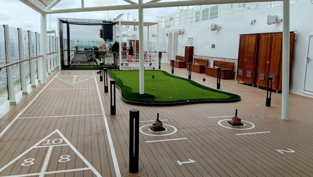The image shows the sports deck on Cunard's Queen Anne cruise ship, featuring a variety of leisure activities. There is a small putting green for golf practice, a shuffleboard court, and a deck quoits setup. The area is covered with a partial roof to provide shade, with glass railings offering clear views of the ocean. Wooden benches and lockers line one side, providing seating and storage for equipment. The space is designed for relaxed outdoor activities, with ample room for passengers to enjoy these classic deck games while taking in the sea breeze.