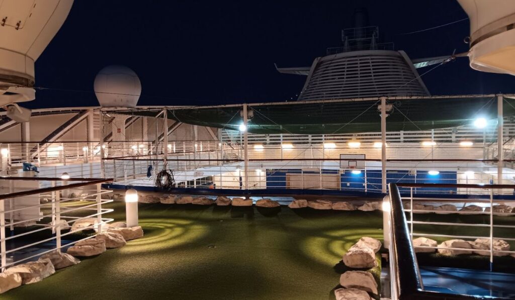 The image shows the sports deck on the Regal Princess at night, featuring a well-lit area with artificial turf, bordered by rocks, possibly part of a mini-golf course. In the background, a netted area suggests spaces for sports activities like basketball or tennis. The scene is serene, illuminated by soft lights, with the ship’s radar dome and funnels visible above the deck.