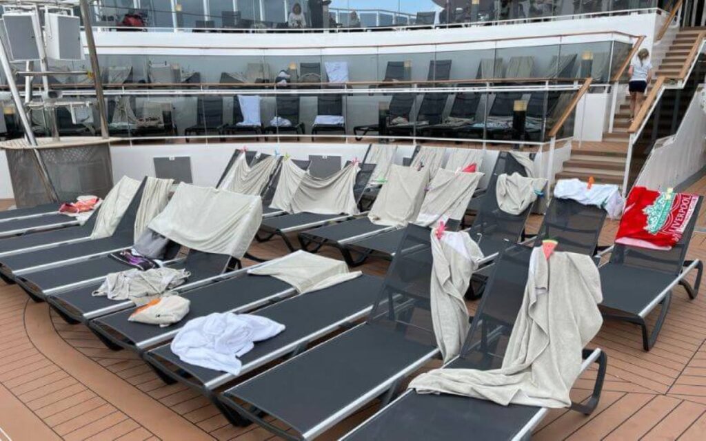 A row of sun loungers on a cruise ship deck, covered with towels and personal items to reserve spots. The loungers are arranged in tiers, allowing for optimal sunbathing and views. Stairs and railings in the background lead to higher deck levels, where additional loungers are also reserved in a similar manner.