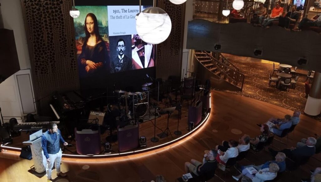 The image shows a talk being held on Cunard's Queen Anne cruise ship, focusing on famous art heists. The presenter is speaking in front of a large screen displaying images related to the 1911 theft of the Mona Lisa from the Louvre, featuring the artwork and a portrait of Vincenzo Peruggia, the man behind the heist. The audience, seated in a cozy, intimate venue, listens attentively. The stage is equipped with musical instruments, hinting at other entertainment events, and the surrounding decor is modern and elegant, with warm lighting and stylish accents.
