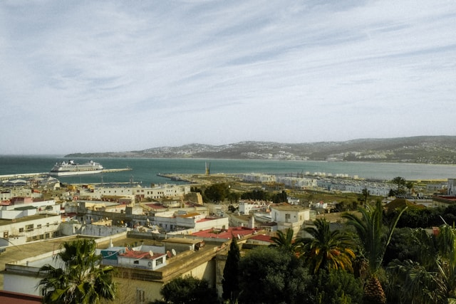 An elevated view of the Port of Tangier in Morocco, showing the harbor with a large cruise ship docked, white buildings of the city's medina, and hills in the distance under a cloudy sky.