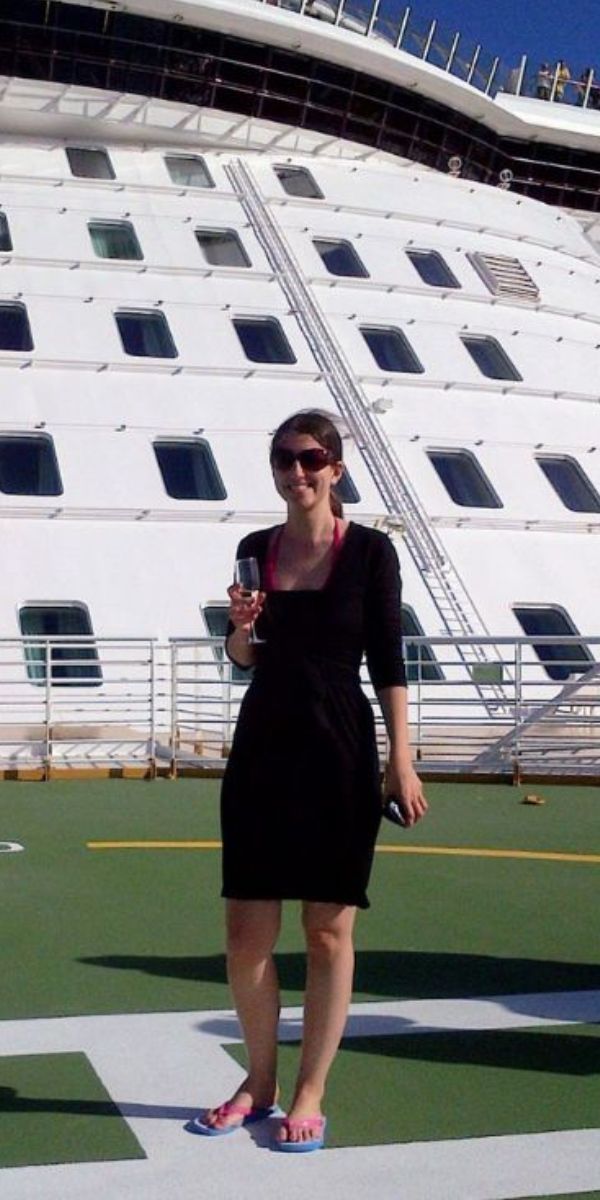 A woman in a black dress and sunglasses stands on the deck of a Royal Caribbean cruise ship, holding a wine glass and smiling, with the multi-storied ship's exterior and clear blue sky in the background.