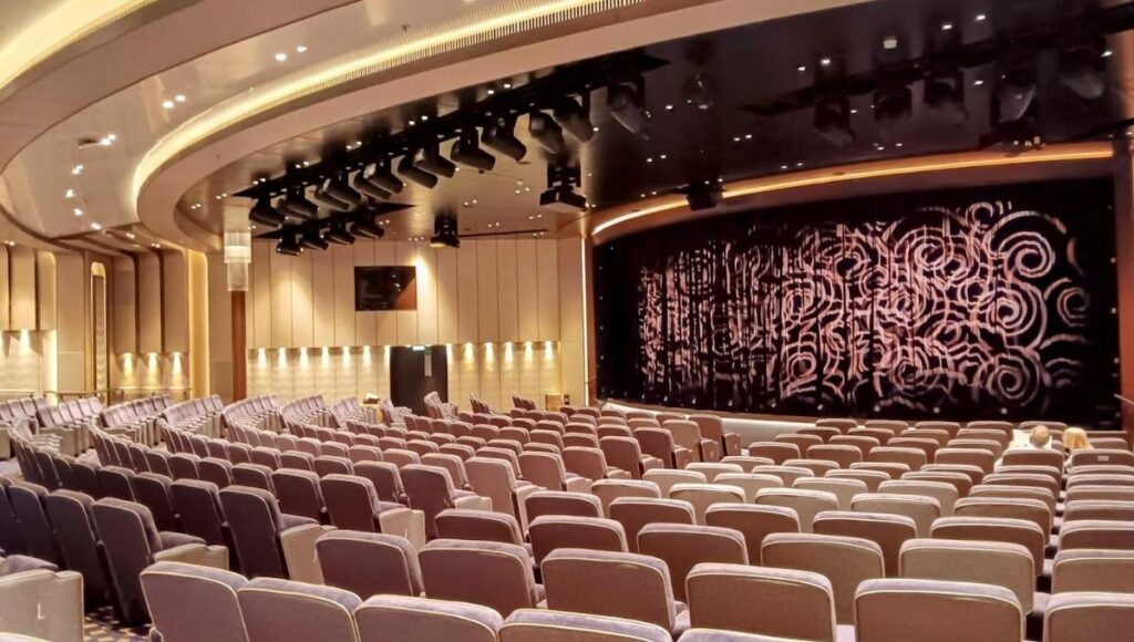 The image shows a luxurious theatre aboard Cunard's Queen Anne cruise ship. The theatre features rows of plush seating arranged to face a large stage with a beautifully patterned curtain. The lighting is soft and sophisticated, highlighting the elegant design of the walls and ceiling, with recessed lighting and a sleek, modern aesthetic. The space is equipped with professional stage lighting, ensuring a premium experience for performances or presentations held in this venue. The setting is comfortable yet upscale, offering an immersive atmosphere for onboard entertainment.