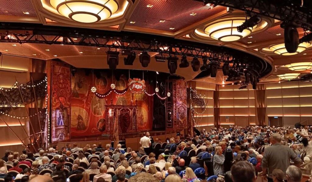 The image shows a full audience inside the theater on the Regal Princess cruise ship. The stage is set for a performance, featuring elaborate decorations with red drapes, lights, and an artistic backdrop in a vintage circus theme. The ceiling has modern circular lights, adding to the warm, inviting atmosphere as passengers wait for the show to begin.