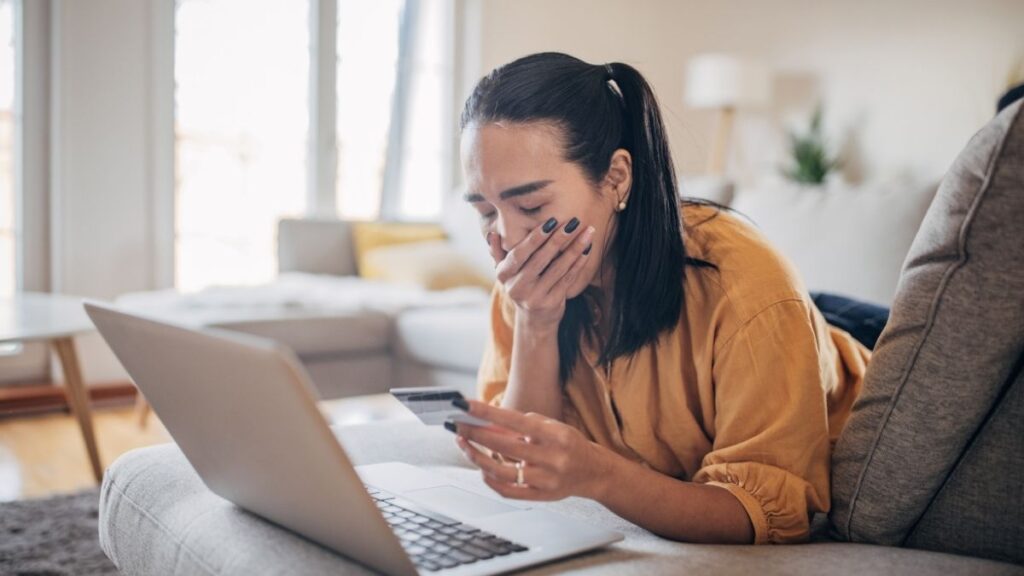 A woman with help hand over her mouth, looking shocked, with a phone in her hand and laptop in front of her.