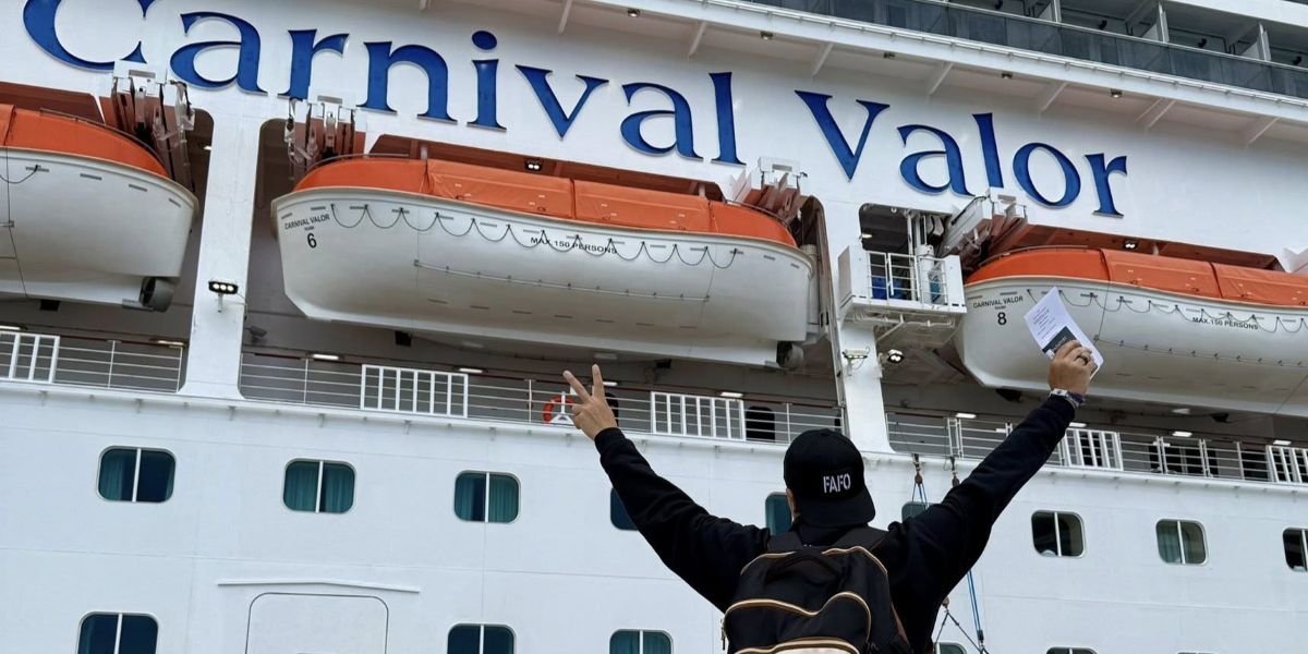 A person wearing a backpack stands on the pier facing the Carnival Valor cruise ship, raising both arms in the air while holding documents in one hand.