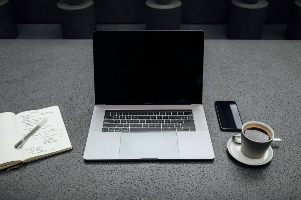 Open laptop on a gray countertop with a notebook and pen on the left, a smartphone on the right, and a cup of coffee on a saucer with a spoon.