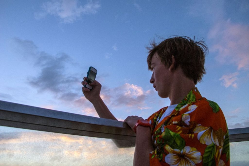 A young person in a vibrant floral shirt leans on the railing of a cruise ship, engrossed in using their smartphone against a dramatic twilight sky.