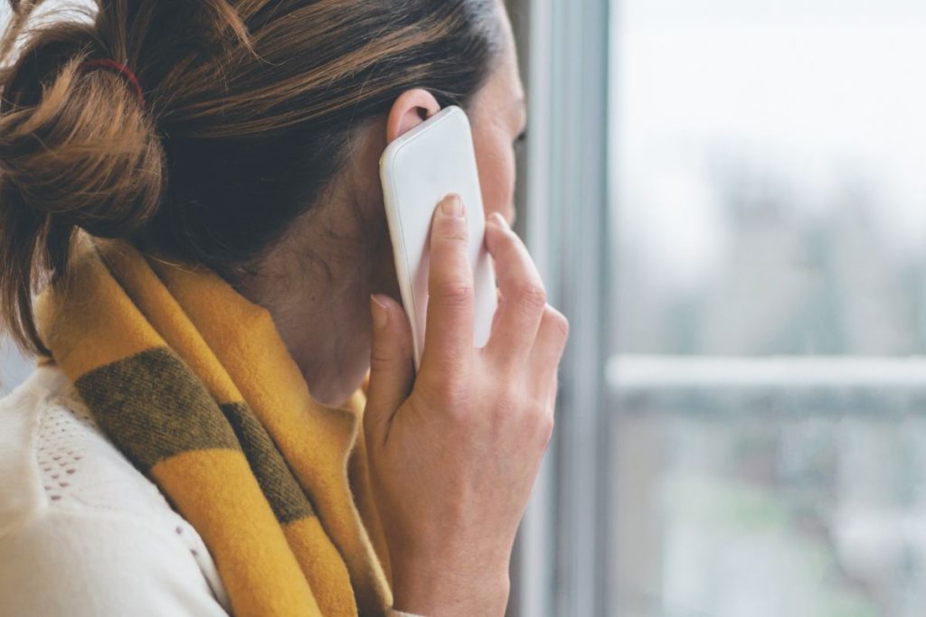 A woman in a cozy yellow scarf is engaged in a conversation on her smartphone, with her back turned to the camera, looking out of a window on a bright day.