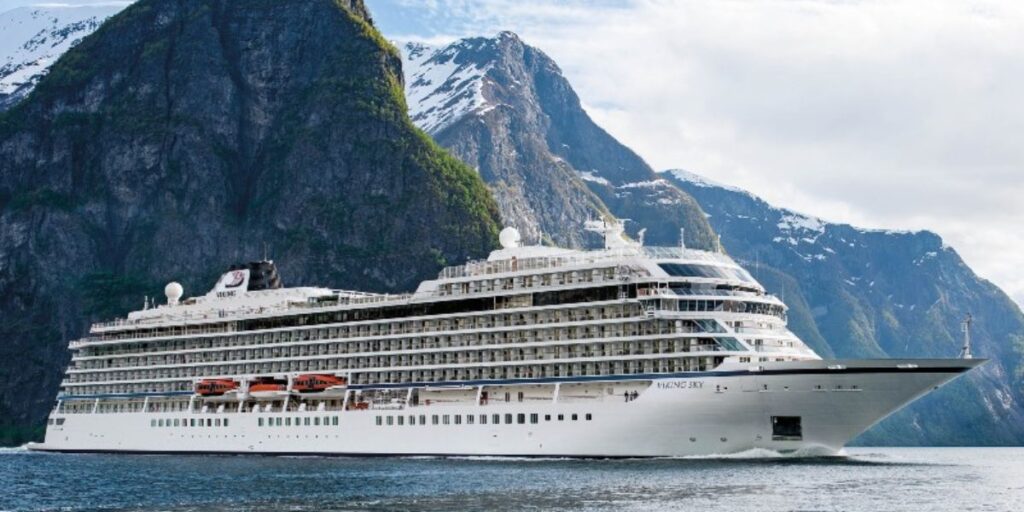 A white Viking cruise ship gracefully navigating through a fjord with steep, lush green cliff faces and snow-capped peaks in the background, under a bright blue sky with scattered clouds.
