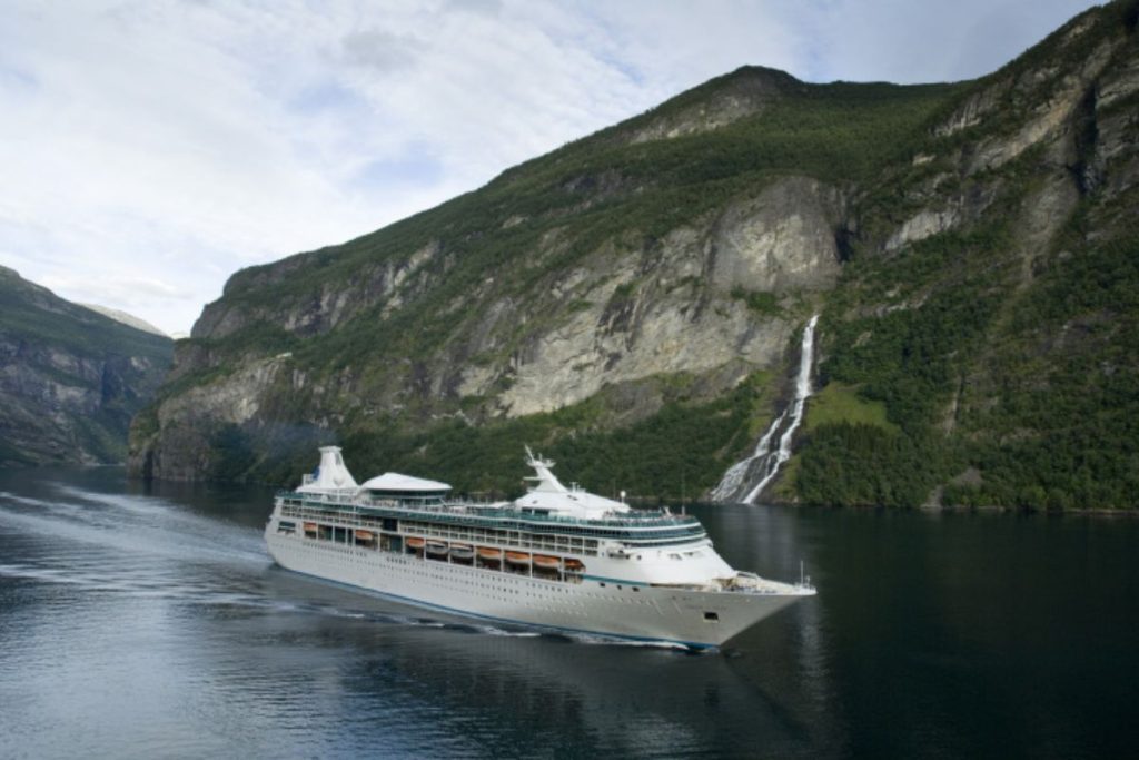 The 'Vision of the Seas' cruise ship glides through calm waters in a fjord, flanked by steep green cliffs and a cascading waterfall, under a partly cloudy sky, evoking a sense of adventure in the Norwegian landscape.