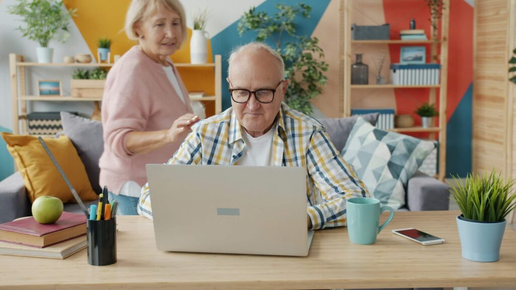 An older couple at home reviewing travel plans on a laptop, with notebooks and a coffee mug on the desk.
