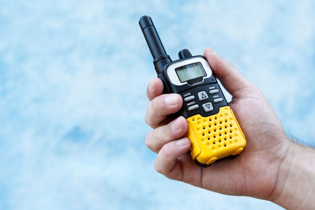 The image shows a handheld walkie-talkie with a black and yellow design. The device has an antenna, buttons for menu and communication functions, and a small screen. It is being held by a person's hand, with a blue, slightly blurred background resembling the sky.