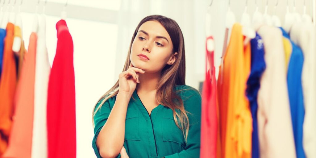 A woman in a green shirt is standing in front of a clothing rack, thoughtfully considering different outfits. The rack is filled with colorful clothing options, including red, orange, blue, and beige garments.