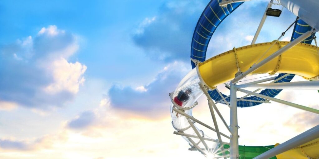 A person speeds through a transparent tube section of a colorful waterslide on a Royal Caribbean cruise ship, with dramatic clouds and blue sky glowing in the background. The twisting yellow and blue slide structure stands tall, creating an adventurous vibe against the scenic ocean sky.