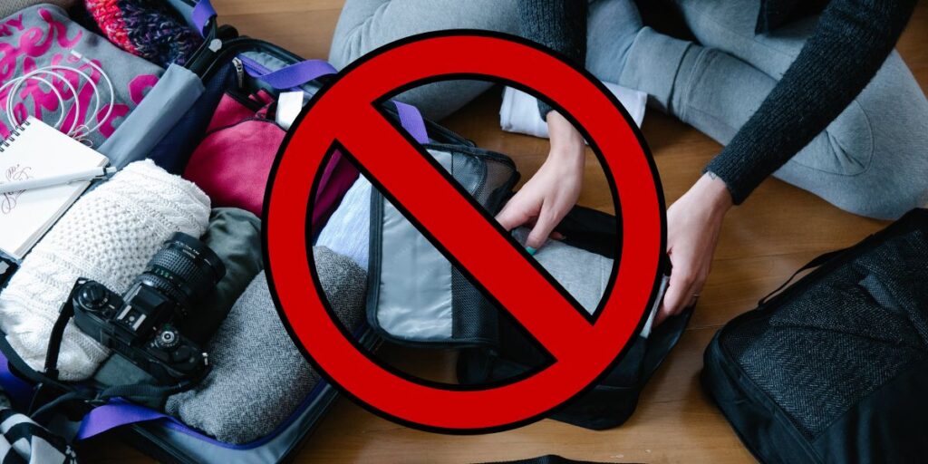 An overhead view of a person's hands organizing a suitcase filled with various travel items, including a camera, clothing, and a notebook, overlaid with a bold red 'no' sign indicating items not to pack or prohibited items for travel.