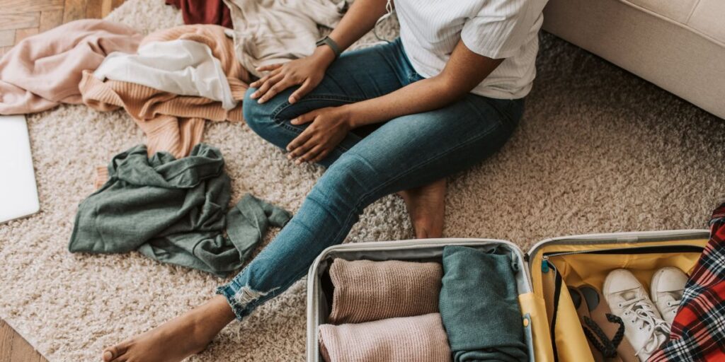 A person sitting on the floor next to an open suitcase, surrounded by a variety of clothes, with a laptop nearby. They appear to be in the process of packing or unpacking, with the emphasis on casual wear and travel essentials.