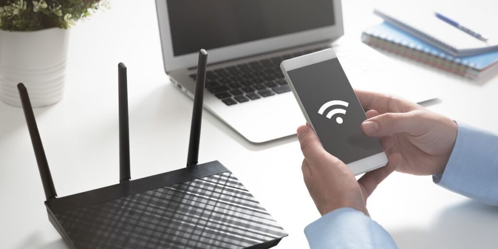 Close-up of hands holding a smartphone with a Wi-Fi signal icon on the screen, indicating connectivity to a wireless router, with a laptop and notepads in the background on a white desk.