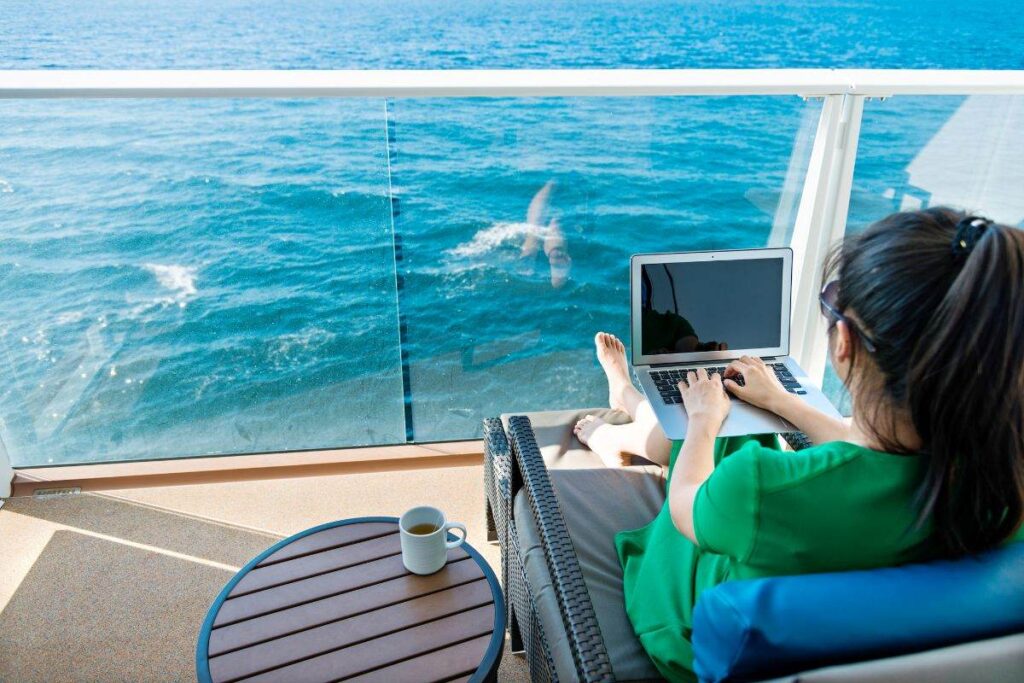 A woman in a green dress is working on her laptop while lounging on a cruise ship balcony. The view showcases the vast ocean, with a small table beside her holding a cup of coffee. She enjoys a serene and productive moment with the sea in the background.