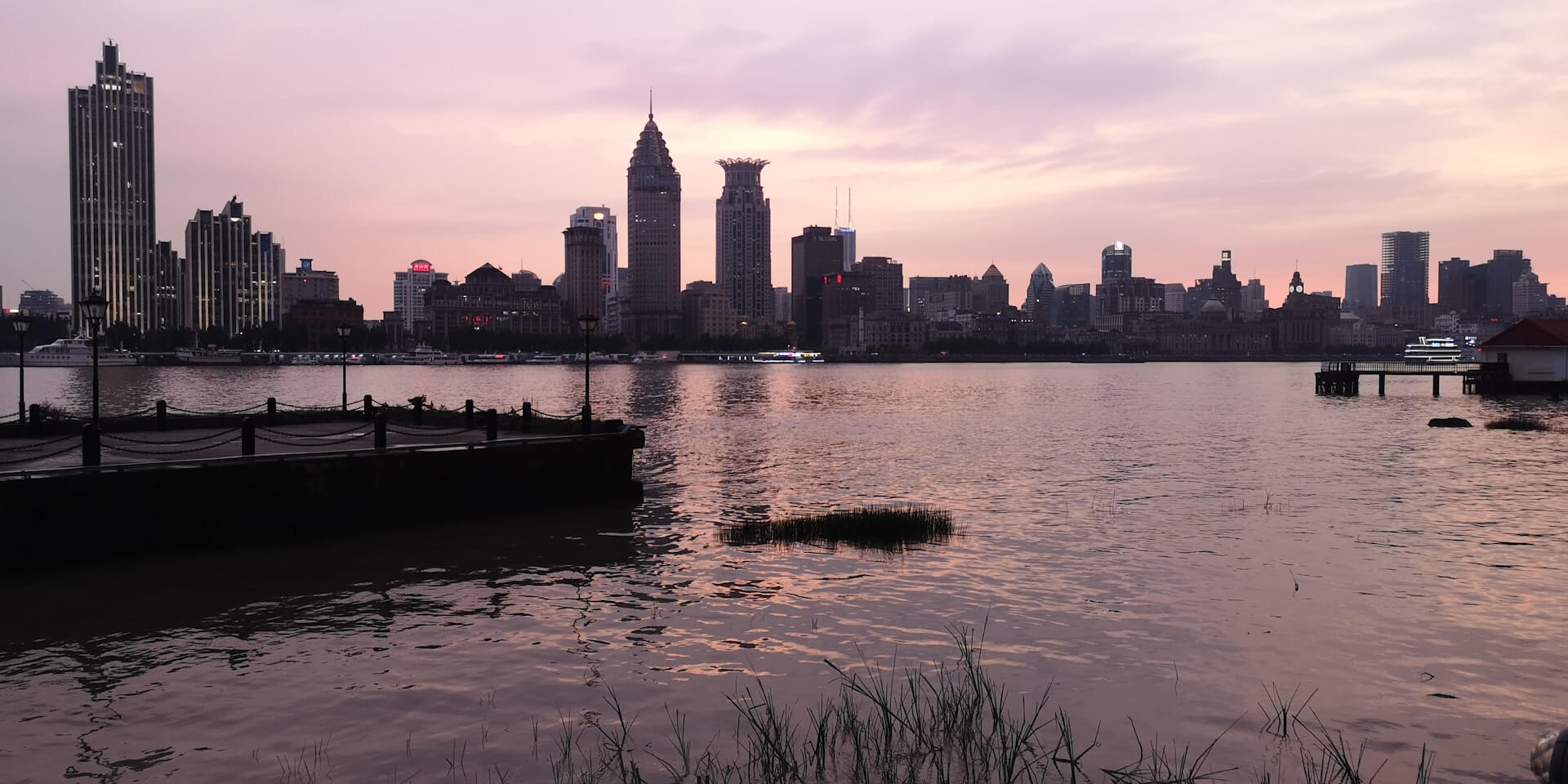 Philadelphia skyline at sunset across the wide Delaware River, with reflections on the water and a soft pink sky.
